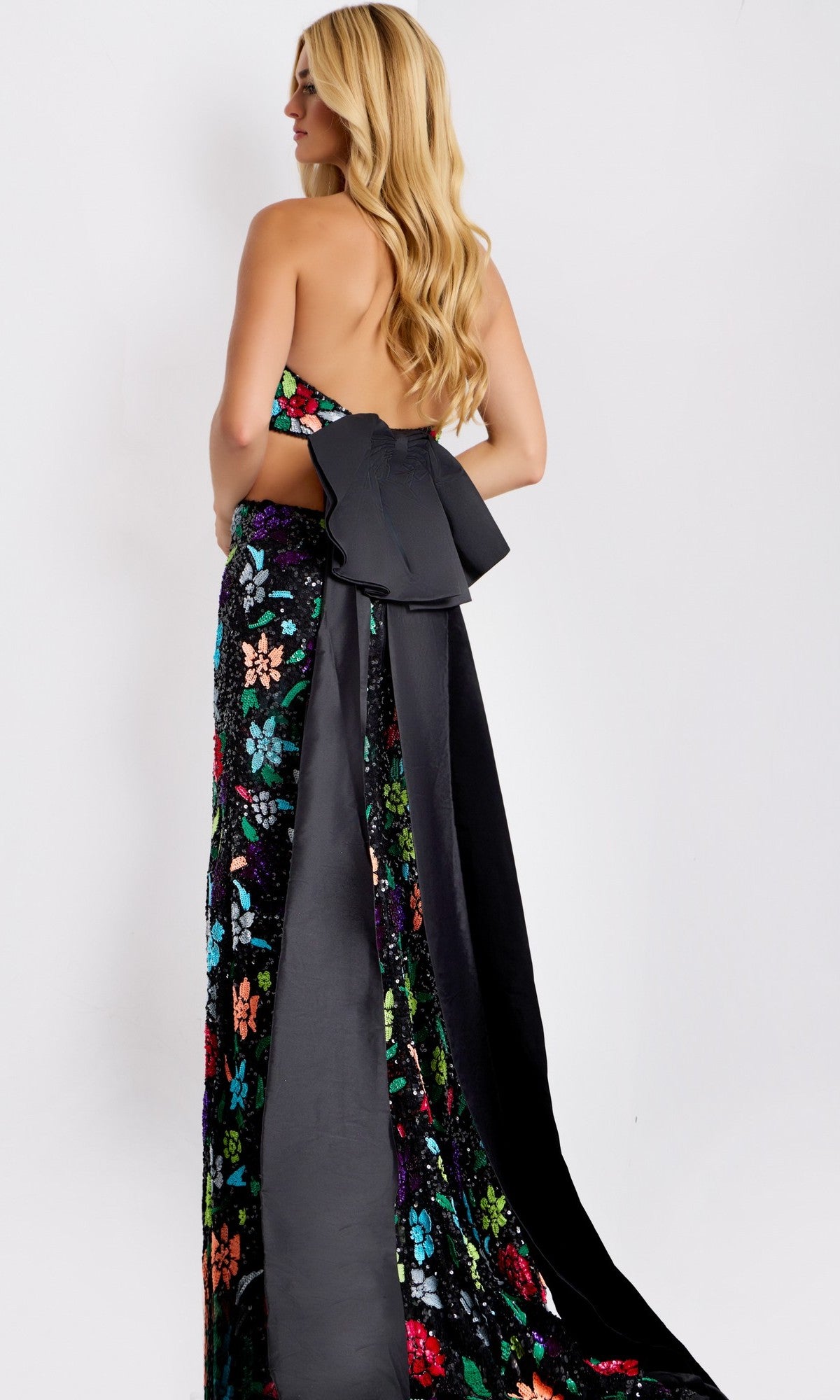A woman models the Jovani 49251 long prom dress with colorful sequins, floral design, open back, halter neckline, and a large black bow detail, standing against a plain white background.